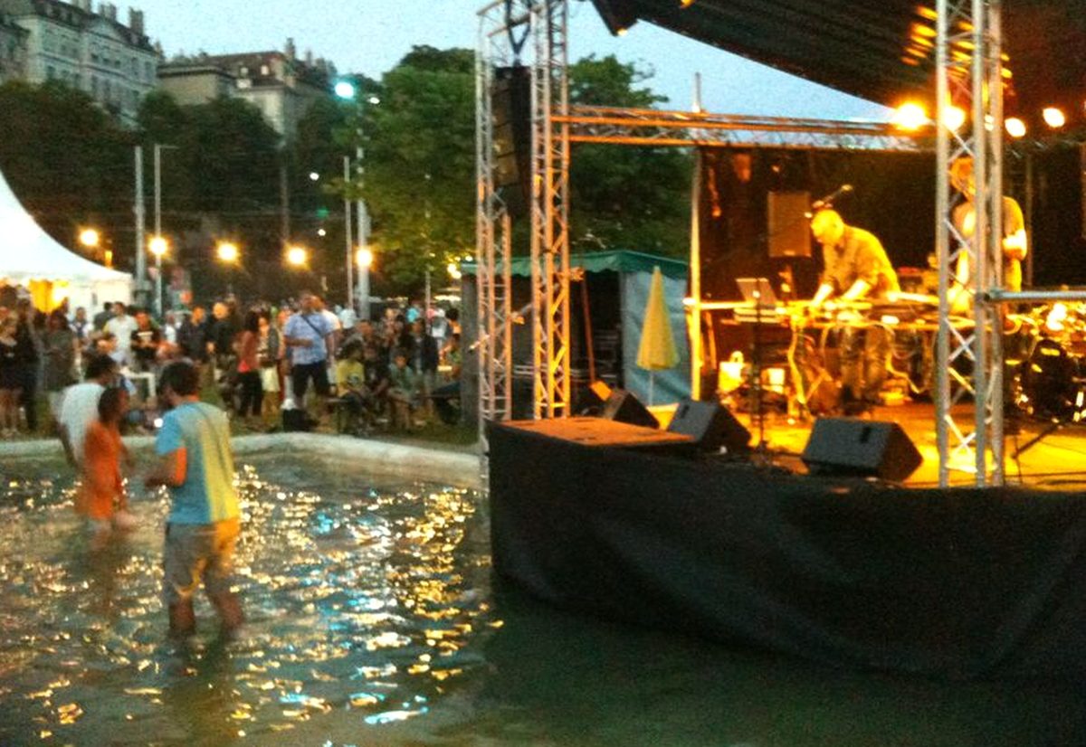 Scène en plein air de la Fête de la musique à Genève au crépuscule, spectateurs les pieds dans un bassin devant le DJ
